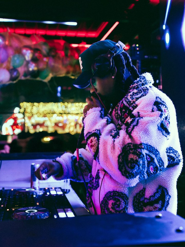 BRODY at the decks in a patterned jacket and headphones, lit in red and blue