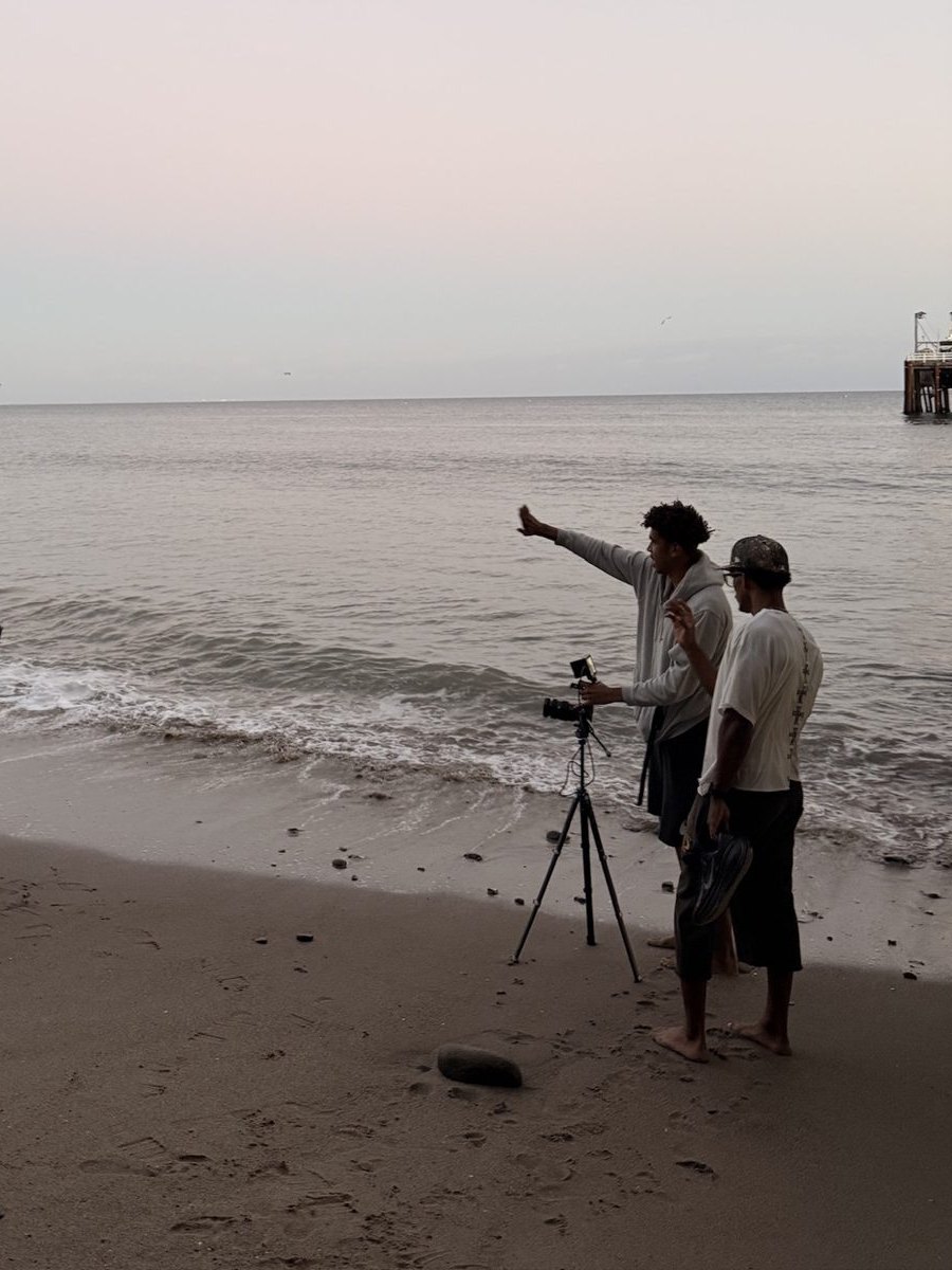 Behind the scenes on the beach shoot for RA-MY.’s “Pacific Ghost .”: two crew at a camera on a tripod at the water’s edge, a pier in the distance